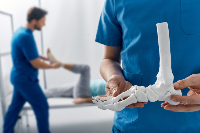 Arzt, der ein anatomisches Modell des Fußskeletts zeigt, während sein Assistent das verletzte Bein eines Patienten untersucht